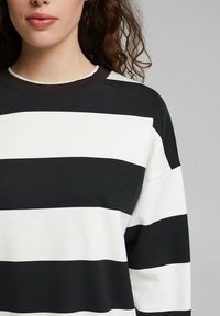 Femme portant un pull à larges rayures horizontales noires et blanches avec un col rond, sur un fond gris.