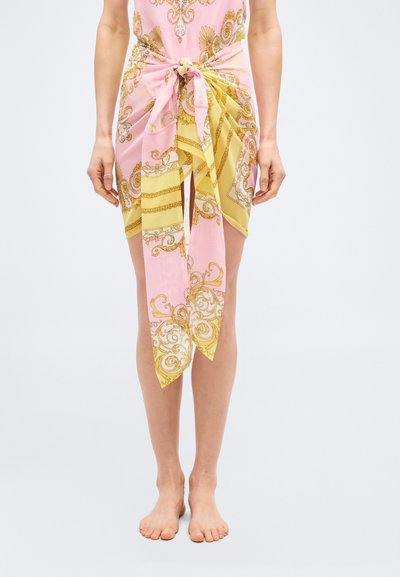 Kvinde iført et pink og gulmønstret tørklæde bundet som en nederdel, stående barfodet mod en ensfarvet lys baggrund.