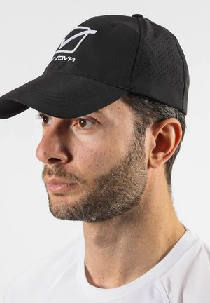 Uomo con capelli corti e scuri e barba, che indossa un cappellino nero traforato con logo bianco e una camicia bianca, che guarda verso sinistra su uno sfondo semplice.