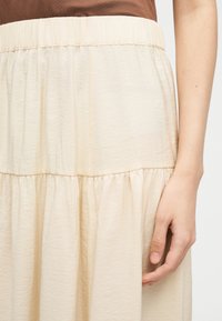 Κοντινό πλάνο ενός ατόμου που φοράει μπεζ φούστα με λάστιχο στη μέση και καφετί κορυφή, με το δεξί του χέρι να πέφτει χαλαρά δίπλα του.