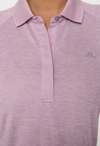 Ingrandimento di una persona che indossa una polo a trama lilla con colletto, due bottoni e un piccolo logo sul lato destro del petto.