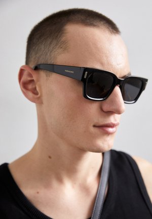 Occhiali da sole rettangolari neri con lenti scure, caratterizzati da un logo "Ferragamo" dorato sul lato e una finitura lucida. Indossati da una persona.