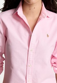 Světle růžová košile na knoflíky s klasickým límcem. Obsahuje malou logo akcentaci na levé části hrudníku a viditelné šicí detaily.