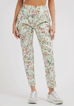Kvinde iført lysegrønne bukser med blomsterprint og hvide sneakers, stående foran en ensfarvet baggrund, hænder hviler langs siden.