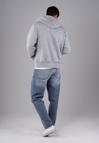 Man wearing a gray hoodie, blue jeans, and white sneakers standing with back facing the camera against a plain gray background.