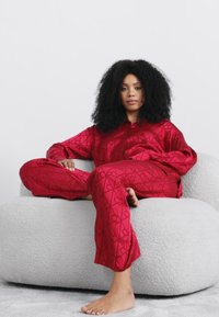 Set de pijamale din satin roșu cu model cu inimioare, având un top cu nasturi și pantaloni cu picior drept, purtat în timp ce stă pe un scaun texturat, de culoare gri deschis.