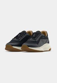 Mustade ja tumehallide paeltega tossude paar, millel on valged tallad ja pruunid välistallad, kuvatud valgel taustal.