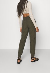 Olivgrüne Jogginghosen mit elastischem Bund und gerippten Beinabschlüssen, kombiniert mit einem langärmligen beigefarbenen Oberteil und schwarzen Sandalen mit Riemchen.