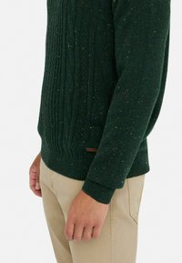 Grüner Strickpullover mit strukturiertem Kabeldesign, mehrfarbigen Flecken, gerippten Bündchen und Saum, kombiniert mit hellbeigen Hosen.