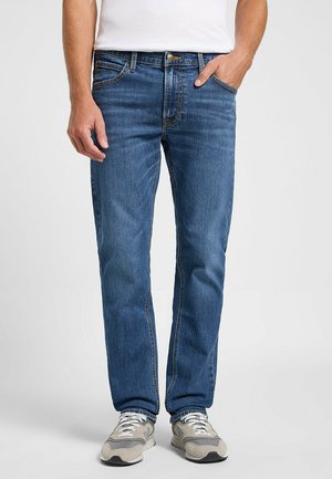 Jeans in denim blu scuro con taglio dritto, dotati di chiusura zip, cinque tasche e cuciture a vista. Indossati con sneakers grigie.