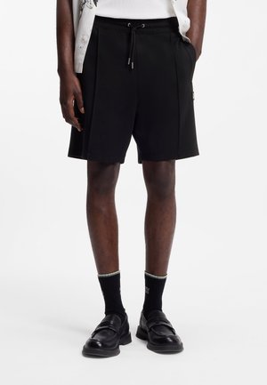 Mand iført sorte shorts med snøre, sorte sokker og sorte loafers, stående med den ene hånd i lommen mod en hvid baggrund.