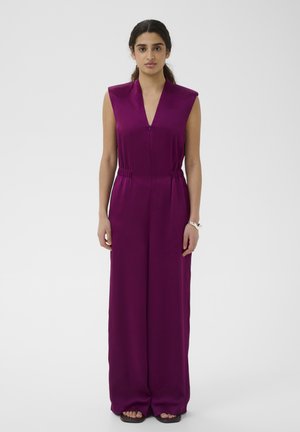 Žena s tmavými vlasmi v bezrukávovom hlbokofialovom overal s širokými nohavicami stojaca na jednoduchom svetlom pozadí.