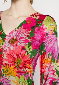Femme portant une robe florale multicolore avec de grands motifs floraux roses, rouges, orange et verts, à décolleté en V, vue du cou à la taille.