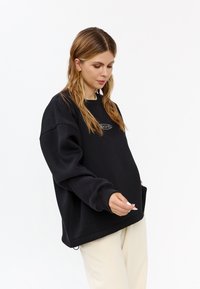 Felpa nera oversize con scollo rotondo, polsini a costine e logo ricamato sul petto. Abbinata a pantaloni chiari.