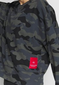Sweat-shirt camouflage dans des tons de gris et noir. Étiquette rouge Calvin Klein Performance sur l'ourlet. Tissu doux, coupe décontractée.