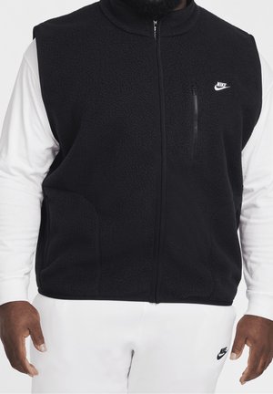 Homme portant une veste polaire noire zippée sur une chemise blanche à manches longues et un pantalon de survêtement blanc Nike avec des logos sur la veste et le pantalon.