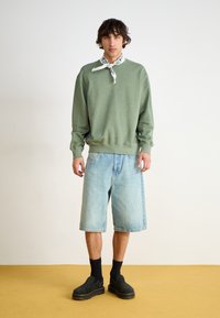 Green sweatshirt, denim shorts, black boots. Sweatshirt has ribbed cuffs and hem; shorts are loose-fitting, reaching mid-thigh. White bandana accent.