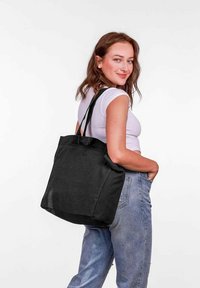 Černá plátěná taška s dvěma ramenními popruhy, čtvercového tvaru, minimalistického designu a hladké textury, kterou drží osoba v bílém topu.