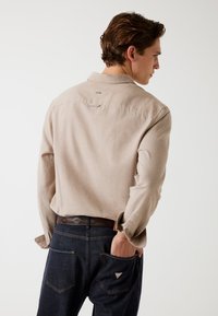 Beige overhemd met lange mouwen en een kraag, met een subtiele logodetail op de achterkant, gecombineerd met donkere denim jeans met een driehoeklogo op de zak.