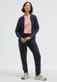 Dunkelblaue Zip-Jacke über einem hellrosa T-Shirt, kombiniert mit dunkelblauen Jogginghosen. Weiße und silberne Sneakers runden den lässigen Look ab.