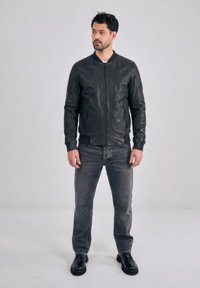 Man staat tegen een effen achtergrond gekleed in een zwart leren jack, vervaagde zwarte spijkerbroek en zwarte schoenen, kijkt naar de zijkant met ontspannen handen.