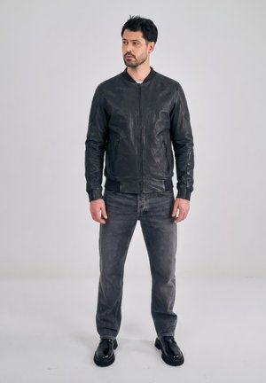 Uomo in piedi davanti a uno sfondo semplice, indossa una giacca di pelle nera, jeans neri sbiaditi e scarpe nere, guarda di lato con le mani rilassate.