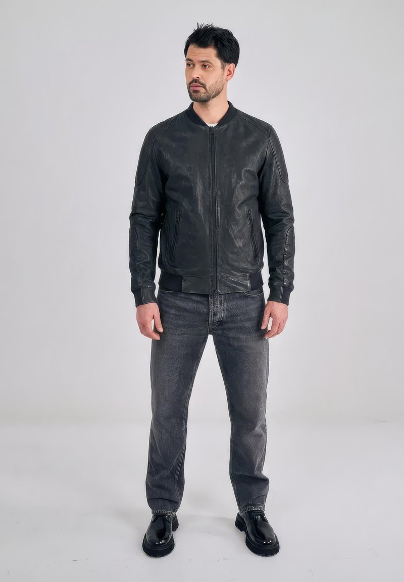 Man staat tegen een effen achtergrond gekleed in een zwart leren jack, vervaagde zwarte spijkerbroek en zwarte schoenen, kijkt naar de zijkant met ontspannen handen.