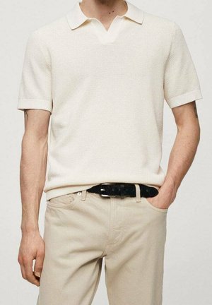 Homme portant un polo à manches courtes blanches texturées, rentré dans un pantalon beige clair avec une ceinture noire, main dans la poche.