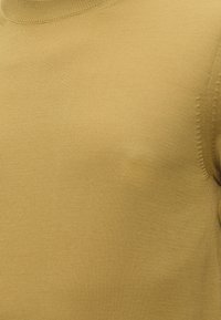 Κοντινή λήψη υφάσματος πλεκτού πουλόβερ μουσταρδί με λεπτή, λεία υφή και διακριτική ριμπ ζώνη κοντά στη λαιμόκοψη και τη ραφή του ώμου.