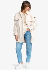 Kremfarget fleecejakke med to frontlommer og knapplukking, båret over en lys grafisk T-skjorte, kombinert med lyseblå jeans og hvite joggesko.