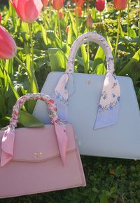 Twee pastelkleurige handtassen met handvatten gewikkeld in een bloemensjaal rusten tussen groene bladeren en roze tulpen in het zonlicht.