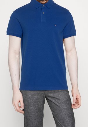 Niebieska koszulka polo z klasycznym kołnierzykiem, krótkimi rękawami i małym logo na piersi; zestawiona z szarymi spodniami w wzór.