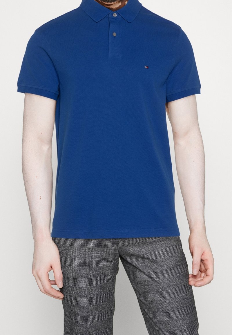 Chemise polo bleue avec un col classique, des manches courtes et un petit logo sur la poitrine ; associée à un pantalon gris à motifs.