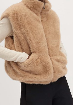 Beige pelsvest uden ærmer med lynlås, båret over en hvid langærmet top, hænderne hvilende i vestens lommer, parret med sorte bukser.
