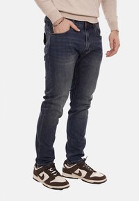 Jeans in denim blu scuro, slim fit con leggeri effetti di sbiadimento, cinque tasche, indossati con sneakers marroni e bianche. Tessuto testurizzato, design classico.