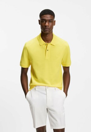 Mann steht mit den Händen in den Taschen, trägt ein hellgelbes Poloshirt und weiße Shorts vor einem schlichten weißen Hintergrund.