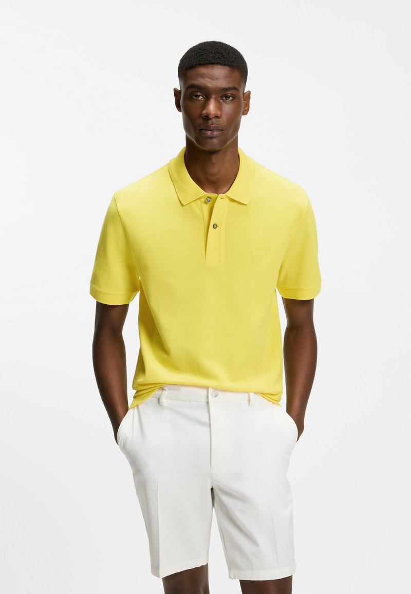 Un homme se tient les mains dans les poches, portant un polo jaune vif et un short blanc, devant un fond blanc uni.