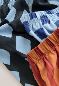 Gros plan de trois bandes élastiques sur un tissu à motifs : noir avec des losanges bleus, blanc avec des formes abstraites bleues et orange avec des rayures rouges.