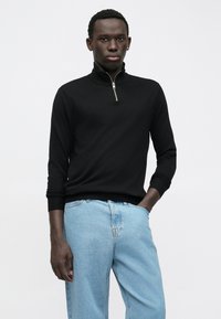 Mlad moški v črnem puloverju z zadrgo in svetlo modrih kavbojkah, ki stoji s svojo roko v žepu nasproti enostavne bele ozadja.