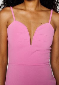 Roze jurk met een diepe V-hals en dunne bandjes. Glad materiaal met subtiele plooien in de buurt van de halslijn en een aansluitende pasvorm.