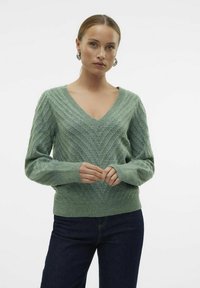 Maglione verde a V in maglia con motivo a zigzag testurizzato, maniche lunghe, polsini e orlo a coste, abbinato a jeans scuri.
