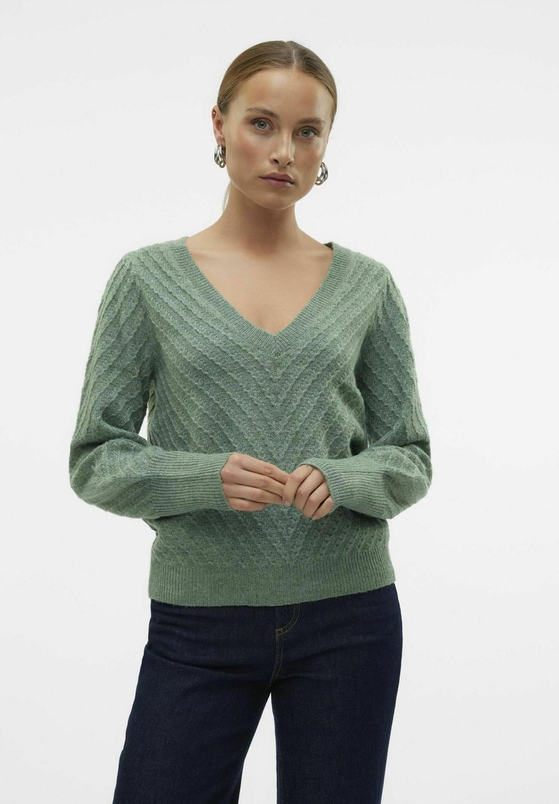 Maglione verde a V in maglia con motivo a zigzag testurizzato, maniche lunghe, polsini e orlo a coste, abbinato a jeans scuri.