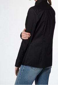 Femme portant un haut à manches longues noir et un jean bleu, tenant son bras gauche avec sa main droite, tournée dos à l'appareil photo.