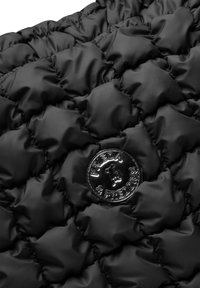 Zwart gewatteerd stof met een puffy textuur. Bevat een ronde, glanzende logo badge met "FRIEDA & FREDDIES" in het midden gegraveerd.
