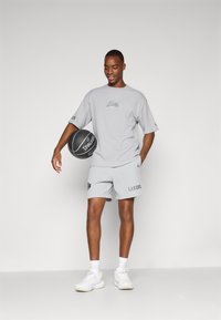 Siva atletska majica i šorts u kombinaciji s natpisom "LAKERS", uz bijele tenisice. Drži crnu košarkašku loptu. Jednostavan dizajn, ležeran kroj.