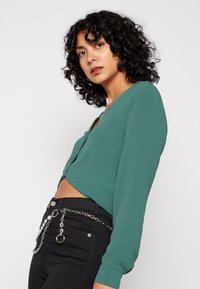 Blusa cropped verde-água com decote em V, mangas longas e um acabamento ligeiramente texturizado, usada sobre jeans pretos com um acessório de corrente na cintura.
