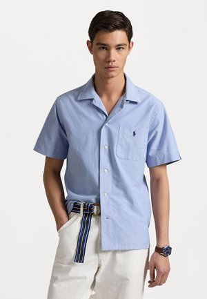 Light blue short-sleeve shirt in a cotton blend, featuring a button-up design, chest pocket, and a small embroidered logo. Paired with white pants and a striped belt.