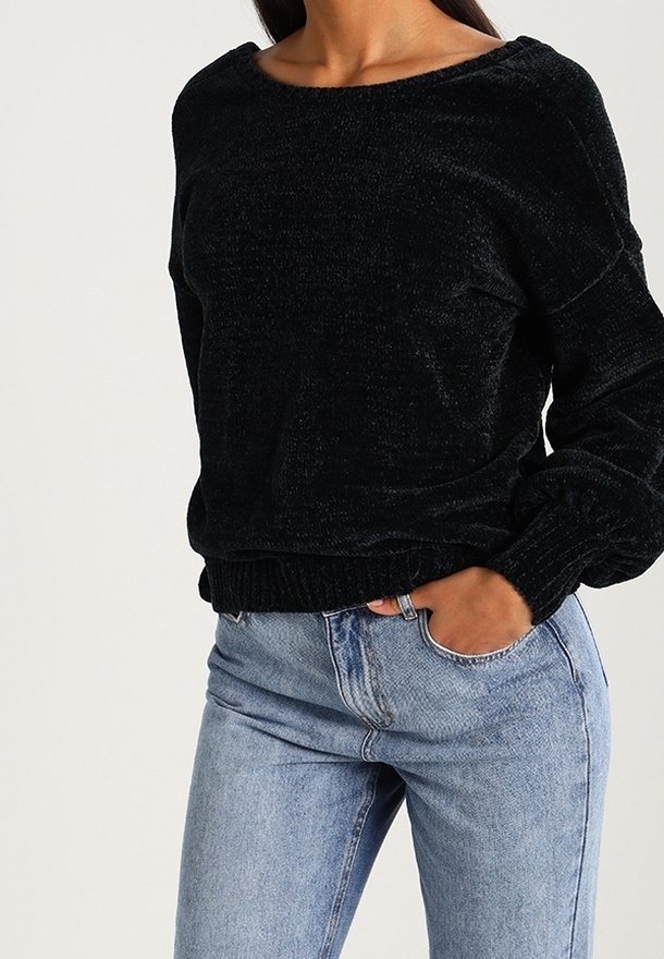 Femme portant un pull noir texturé à manches longues et un jean taille haute bleu clair, une main dans la poche.