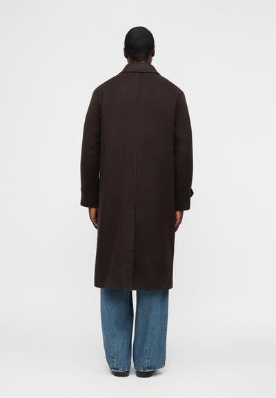 Άνδρας με την πλάτη, φοράει μακρύ σκούρο καφέ παλτό πάνω από άνετο μπλε τζιν και μαύρα παπούτσια, μπροστά σε απλό λευκό φόντο.