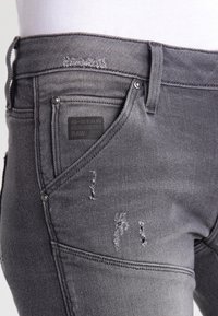 Šedé džíny s úzkým střihem, se sepranými detaily, kovovými prvky a značkovým štítkem na zadní kapse.